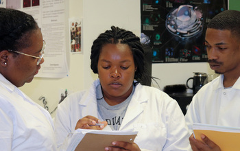 A researcher talks with two students in a laboratory settings. All are wearing lab coats.